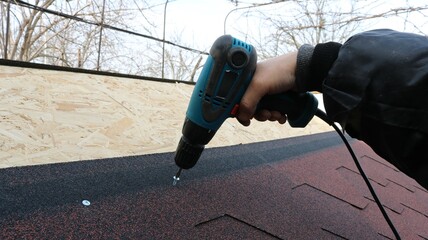 a hand in warm work clothes with a blue electric screwdriver screws fasteners into the roof structure of a low building, connecting soft tiles and an OSB sheet © Skyliz