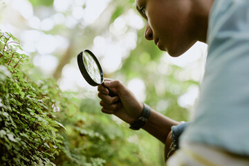 Low angle view closeup of curious African American teen boy studying plants with use of magnifier,...