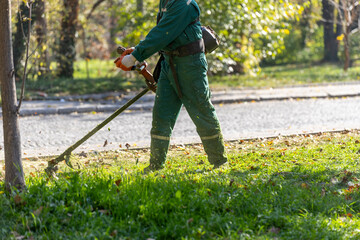 Lawn care worker trimming grass in a park on a sunny day during autumn