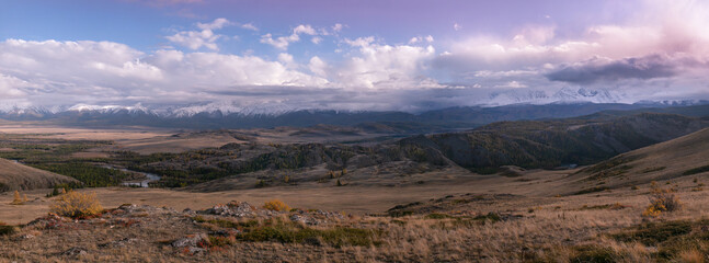 Wide panorama of Chuya steppe in the twilight evening: hilly landscape, endless fields with yellow grass, golden autumn in Altai. In the distance is a mountain ridge with snow covered mountain peaks