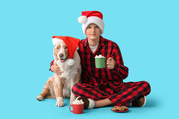 Teenage boy with Australian Shepherd dog in Santa hats holding cup of hot chocolate on blue background