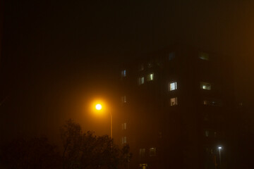 A mysterious and foggy urban scene unfolds, showcasing a tall building adorned with warmly lit...