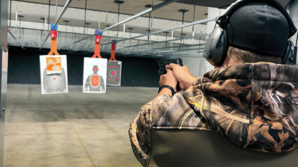 Shooting Practice at Indoor Gun Range. A person wearing camouflage and protective gear takes aim at...
