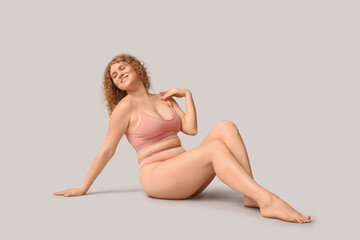 Beautiful young happy body positive woman in trendy underwear sitting on grey background