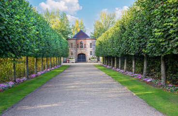 Under blue skies, a tree- and flower-lined crushed stone path leads to a brick garden house on the...