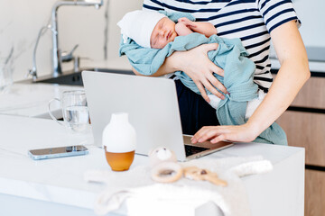 Fototapeta premium Mother multitasking with newborn baby and laptop in kitchen