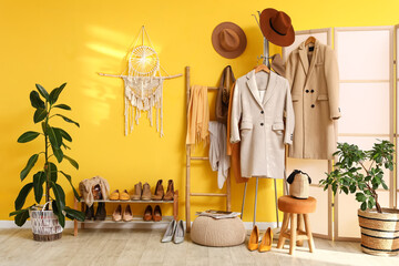 Hanger with clothes and stand with shoes near yellow wall in room