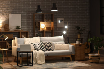 Interior of modern living room with grey sofa, laptop on coffee table and glowing lamps at evening