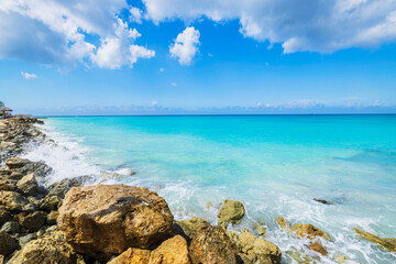 Rocky coastline beach with turquoise waves crashing against rocks under clear blue sky and scattered white clouds. Aruba.