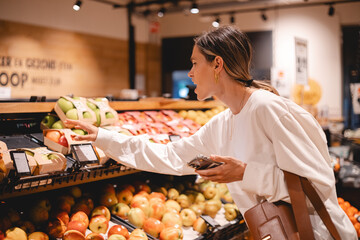 Young woman girl buyer client consumer stands in shop store supermarket choosing food buying puts in basket. Woman choose fruits.