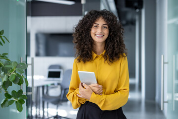 Confident woman in office holding tablet, smiling warmly. Curly hair, yellow blouse, modern...