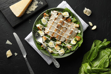 Delicious Caesar salad with chicken on black textured table, flat lay