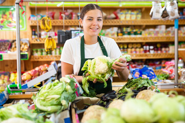 Portrait of young positive european woman working showing fresh cauliflower in grocery store