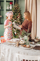 a girl child with mom in the kitchen with a Christmas tree is preparing or cooking ginger cookies for Christmas or New year