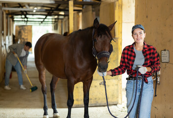 Female owner of horse takes animal out of stall, holds bridle and forces it to follow her. She goes for walk and training with horse, goes out through corridor to racetrack.