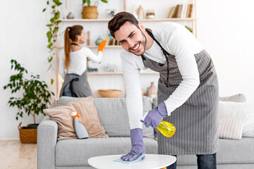 Fototapeta premium Stay at home and cleaning flat spending quarantine together. Cheerful man in apron and rubber gloves with spray and rag wipes dust with wife, near cleaning supplies in interior of living room