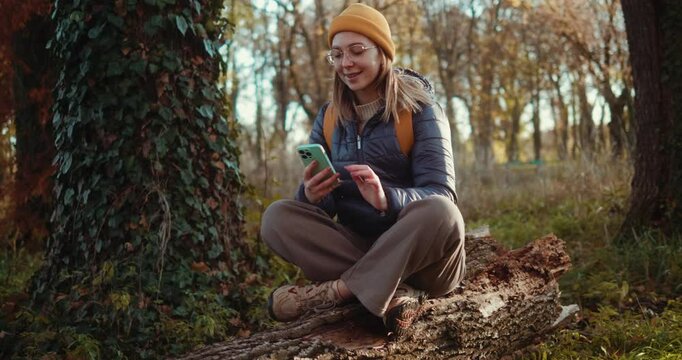 Young adventurer girl sitting on a fallen tree and using a mobile phone, feeling satisfied resting after a trekking walk in the forest