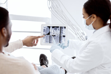 Black female dentist showing xray of teeth treatment process on digital tablet to patient during...