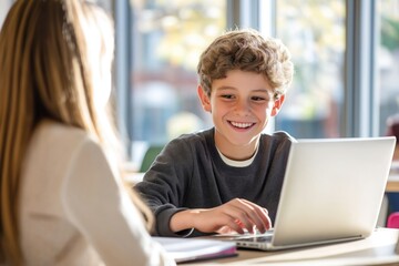Boy smiling happily at laptop. Woman seated nearby. Classroom setting with natural light. Boy actively using laptop. Student engaged with electronic device. Possible tutoring learning session. Casual