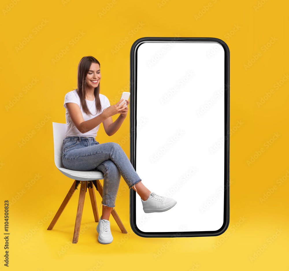 Aufkleber Cheerful smiling female sitting on chair near big cell phone with empty white screen, using mobile device, checking new cool app on orange studio background, mock up for website or application design