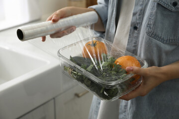Woman putting plastic food wrap over glass container with vegetables in kitchen, closeup