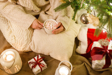 Merry Christmas! Woman in cozy knitted sweater holding warm cup of cocoa with marshmallow under...