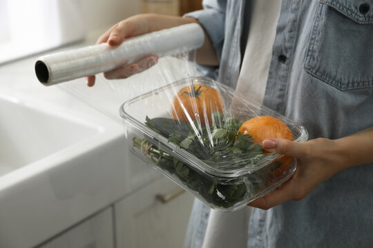 Woman putting plastic food wrap over glass container with vegetables in kitchen, closeup