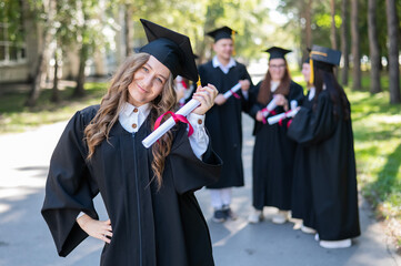 Group of happy students in graduation gowns outdoors. A young girl with a diploma in her hands in the foreground.