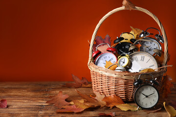 Alarm clocks and dry leaves in wicker basket on wooden table against brown background, space for...
