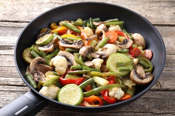 Different vegetables and mushrooms in frying pan on wooden table, closeup