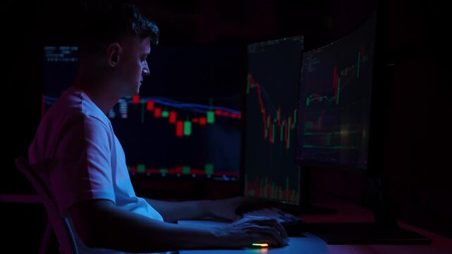 Pensive investor male working from home office as financial analyst, monitoring stock market data on computer screens in dark room, illuminated by colorful glow of charts and graphs, slow motion.