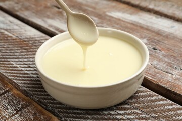 Condensed milk flowing down from spoon into bowl on wooden table, closeup