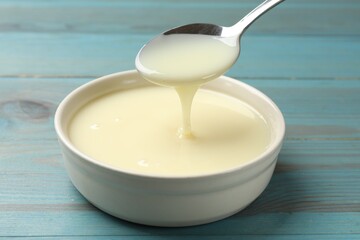 Condensed milk flowing down from spoon into bowl on light blue wooden table, closeup
