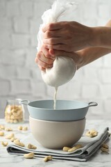 Woman straining cashew milk into colander with bowl at white marble table with nuts indoors, closeup