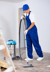 Professional young builder in blue overalls vacuuming floor while cleaning room in building after...