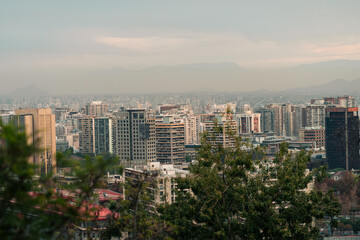 View from the heights of the city of Santiago de Chile. On a cloudy day at sunset, with its iconic long buildings, in the distance, the Andes mountain range, buildings, houses, reflections, windows.