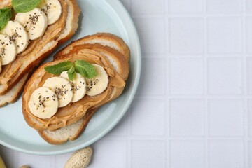 Tasty sandwiches with peanut butter, banana, chia seeds and mint on white tiled table, top view. Space for text