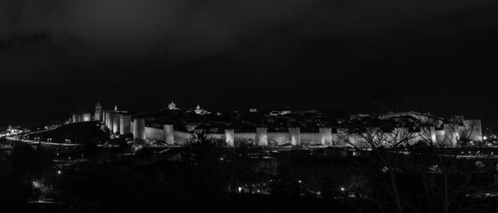 Illuminated nighttime scene showcasing the iconic medieval rampart wall and main entrance gate of &Aacute;vila, Spain. The architecture is beautifully highlighted by soft lighting surrounded by a quiet plaza