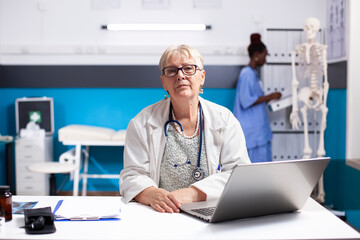 Obraz premium Senior female doctor in lab coat sits at clinic desk with laptop, looking directly at camera. Portrait of elderly physician appearing focused as she prepares for next medical consultation in hospital.
