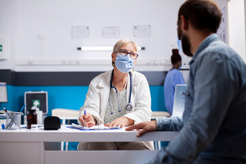 Male patient sharing sickness symptoms with friendly elderly female medic who writes on clipboard during clinic appointment. Senior doctor consults with young man in hospital, both wearing face mask.