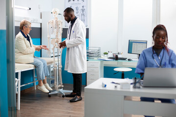 Elderly female patient sits on hospital bed listening attentively to black doctor explaining spinal health as nurse works nearby. Male physician uses skeleton to describe old woman recovery treatment