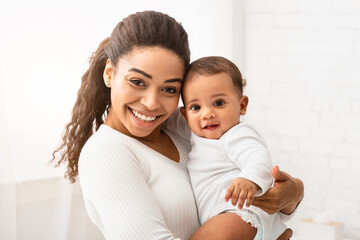 Joyful Black Young Mom Holding And Hugging Adorable Baby Boy Enjoying Motherhood And Child Care Standing In Modern Bedroom At Home, Smiling To Camera. Mother's Day Concept