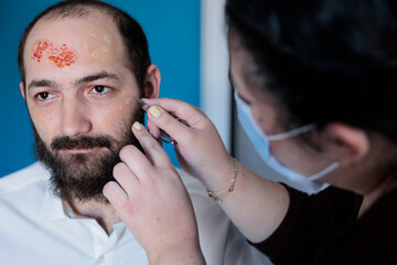 Fototapeta premium Woman peeling actor skin in makeup department after making fake blood wounds, preparing for movie scene. Man getting costumed as undead creeper by makeup artist applying sinister cosmetics