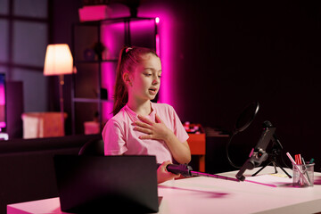 Child utilizes cellphone on selfie stick to shoot introductory segment for vlog. Young influencer in pink neon studio welcomes devoted fan community, filming with mobile phone camera
