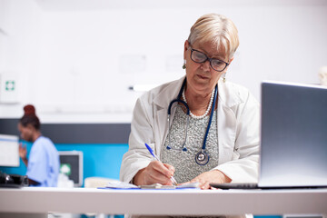 Elderly doctor focused on filling out prescriptions with laptop open for quick access to digital records. Retired female physician seated in clinical office reviewing medical notes on clipboard.