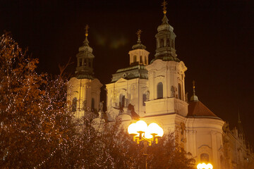 Obraz premium Illuminated historic church towers shine at night, surrounded by vibrant foliage in an urban setting, showcasing intricate architecture and striking lighting that enhance their beauty