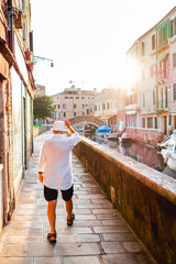 Elderly Man Walking Along a Picturesque Venetian Canal at Sunset Wearing Casual Summer Attire with Sunlit Water and Buildings