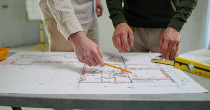 Two men are discussing a house construction project. They are talking standing next to a table in an unadorned room
