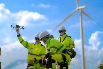 A group of engineers dressed in high-visibility uniforms and helmets conducts an inspection at a wind farm. Using technical devices, they monitor the wind turbines under a bright blue sky.