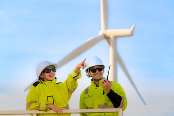 Obraz premium Two engineers in high visibility jackets and hard hats work at a wind energy site, communicating via radio and surveying the surroundings. A wind turbine in the background emphasizes renewable energy.